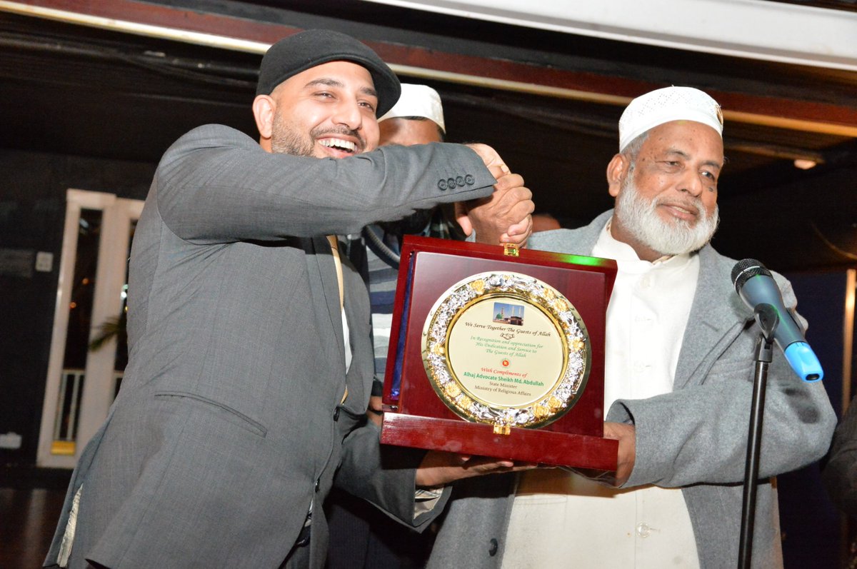 Mohsin Tutla smiling as he receives a plaque that reads We Serve Together The Guests of Allah