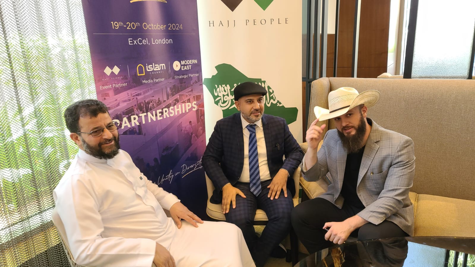 Mohsin Tutla seated with two guests in front of Global Peace and Unity Festival and Hajj People banners