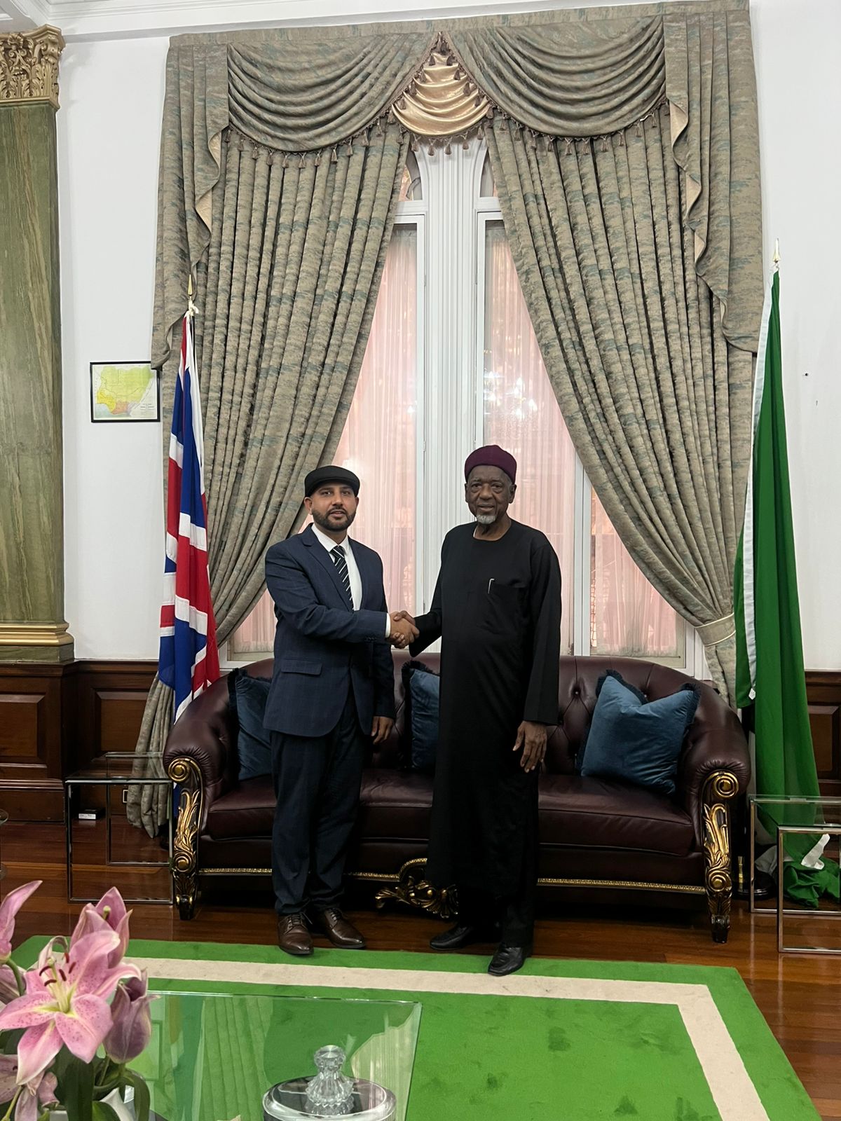 Mohsin Tutla shaking hands with a senior Nigerian dignitary in a formal reception room with British and Nigerian flags
